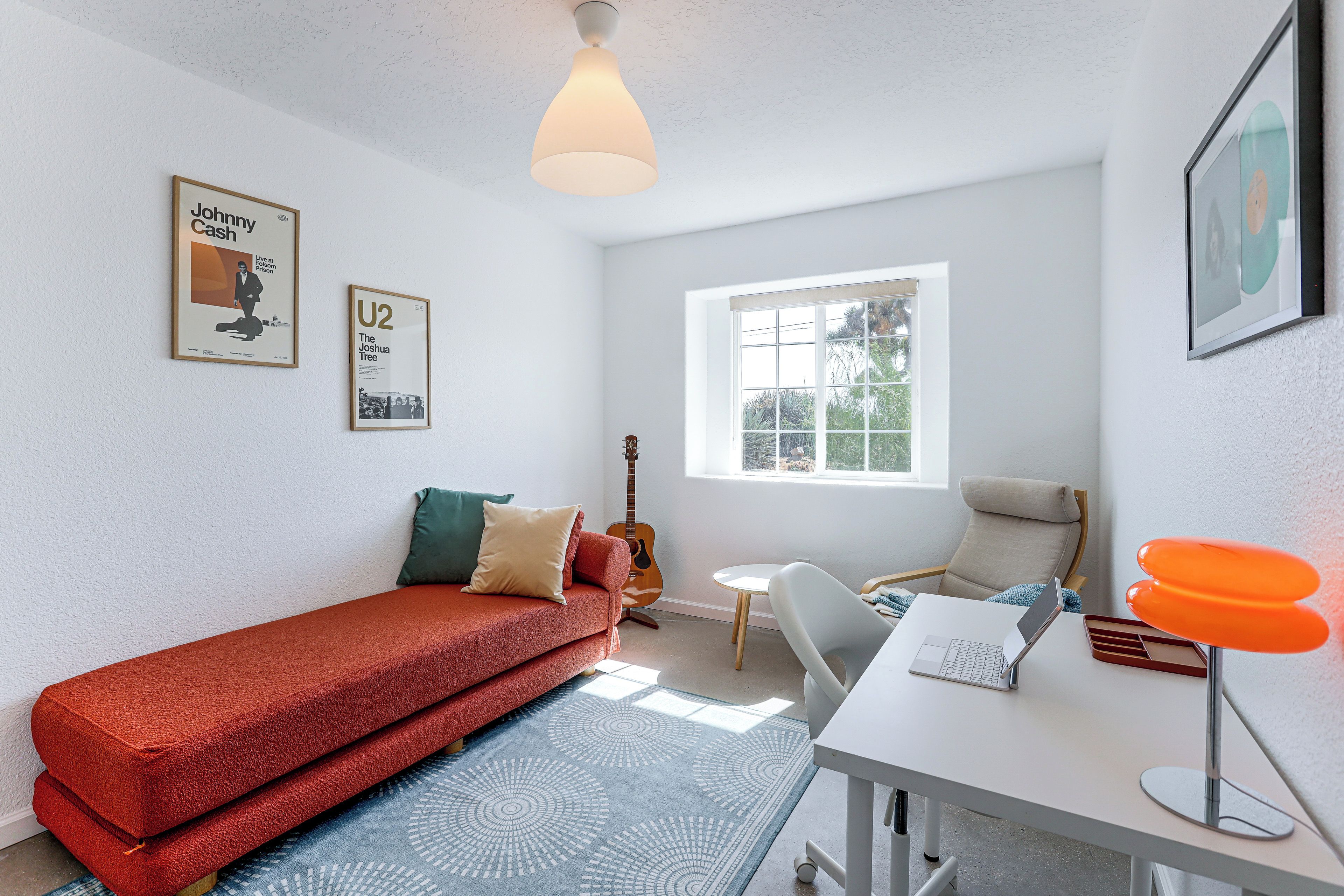A room with a red sofa, a small table with a laptop, a guitar, and a window with a view of trees.