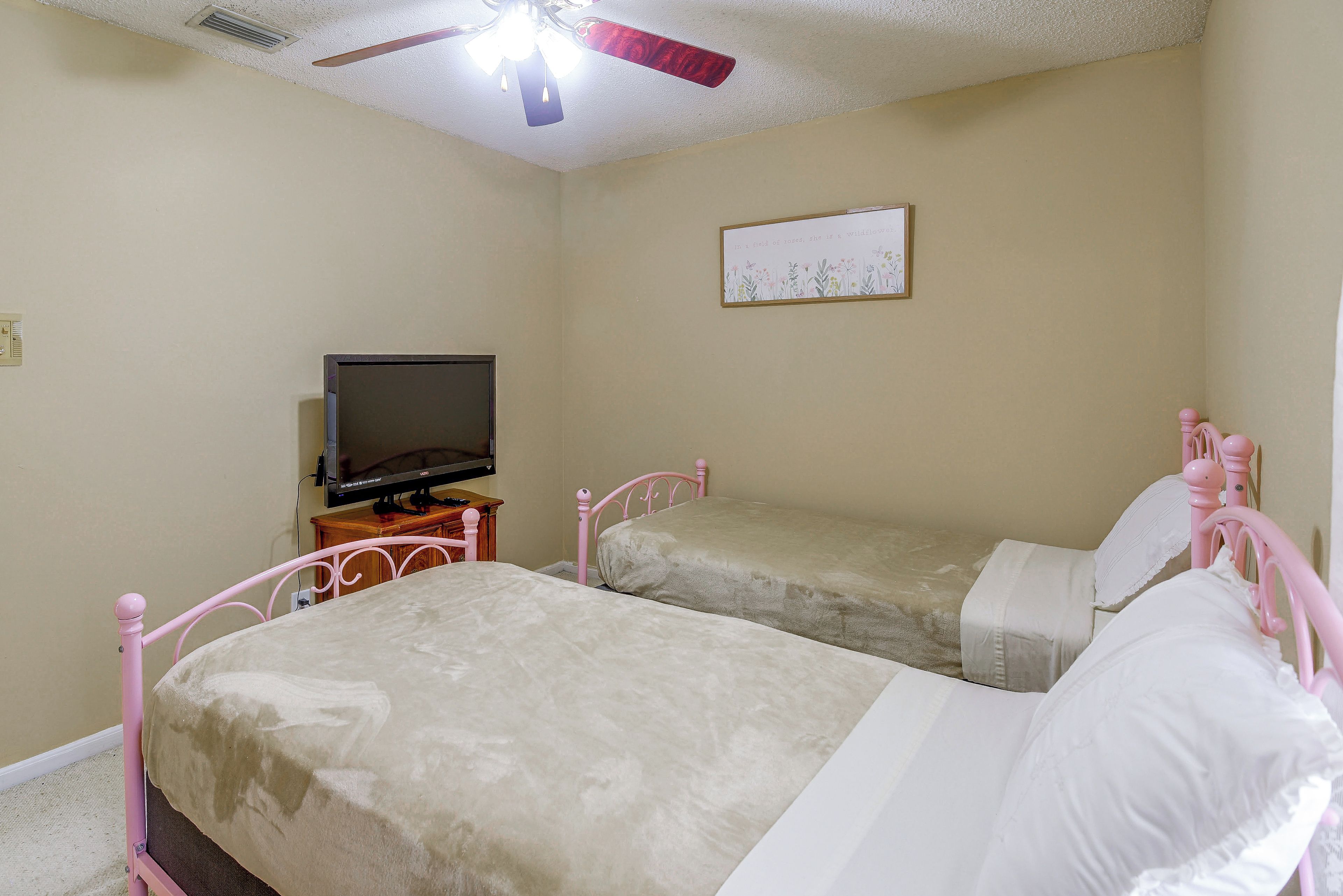 Une chambre avec deux lits, une télévision, un ventilateur de plafond et un tableau accroché au mur.