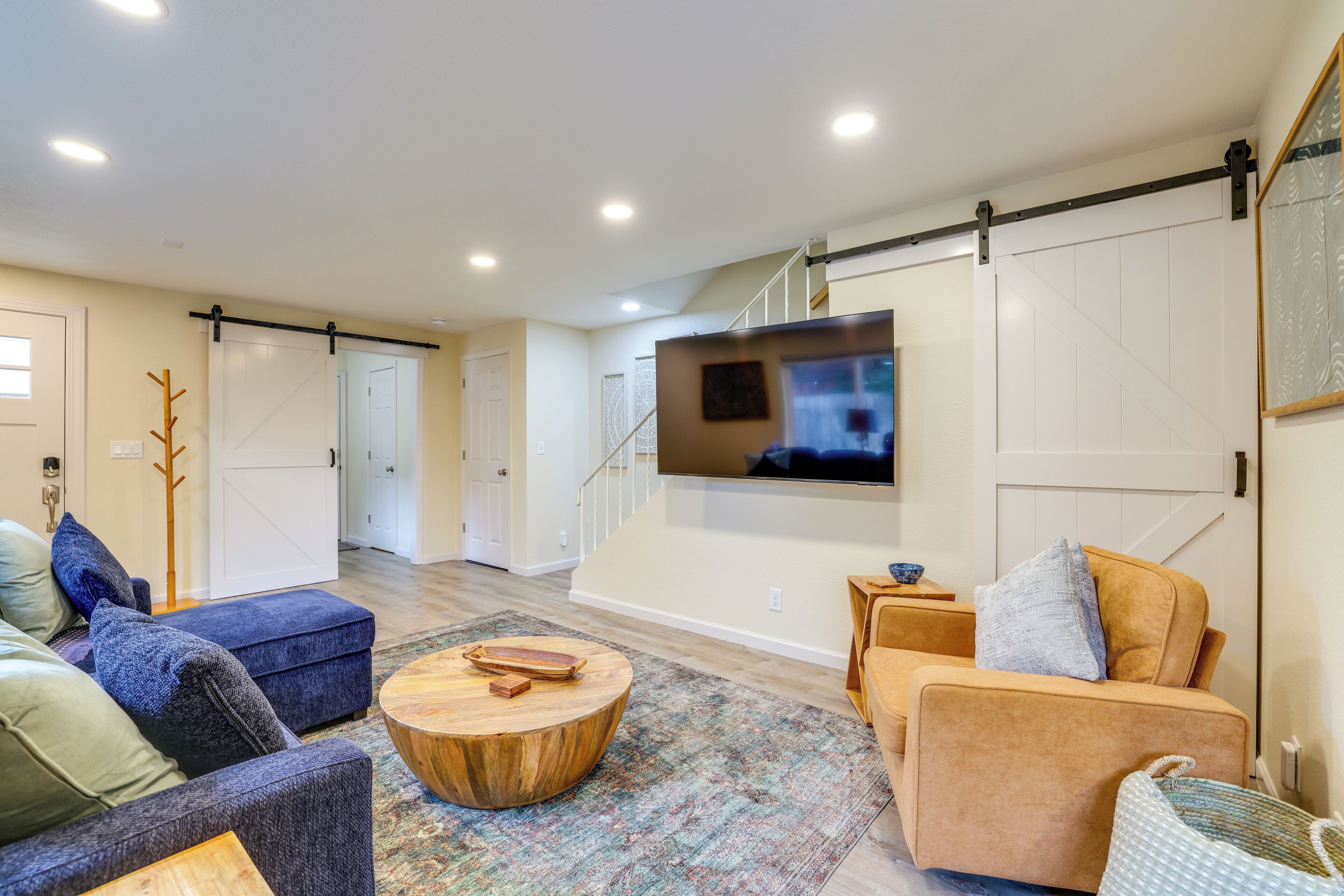 A spacious living room with a large flat-screen TV mounted on the wall, a wooden coffee table, and a variety of seating options including a blue sofa and a tan armchair.