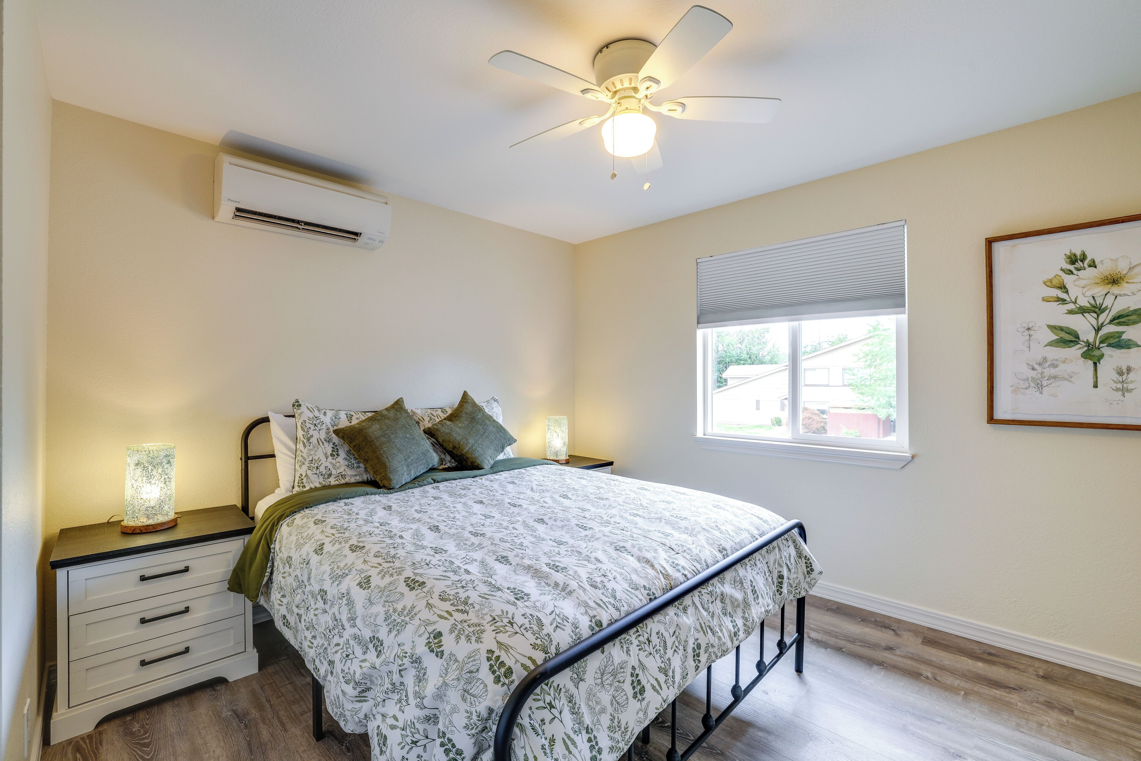 Una camera da letto con un letto, comodini, condizionatore d'aria, ventilatore a soffitto, una finestra con persiane e una stampa botanica incorniciata.