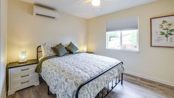 A bedroom with a bed, nightstands, an air conditioner, a ceiling fan, a window with blinds, and a framed botanical illustration.