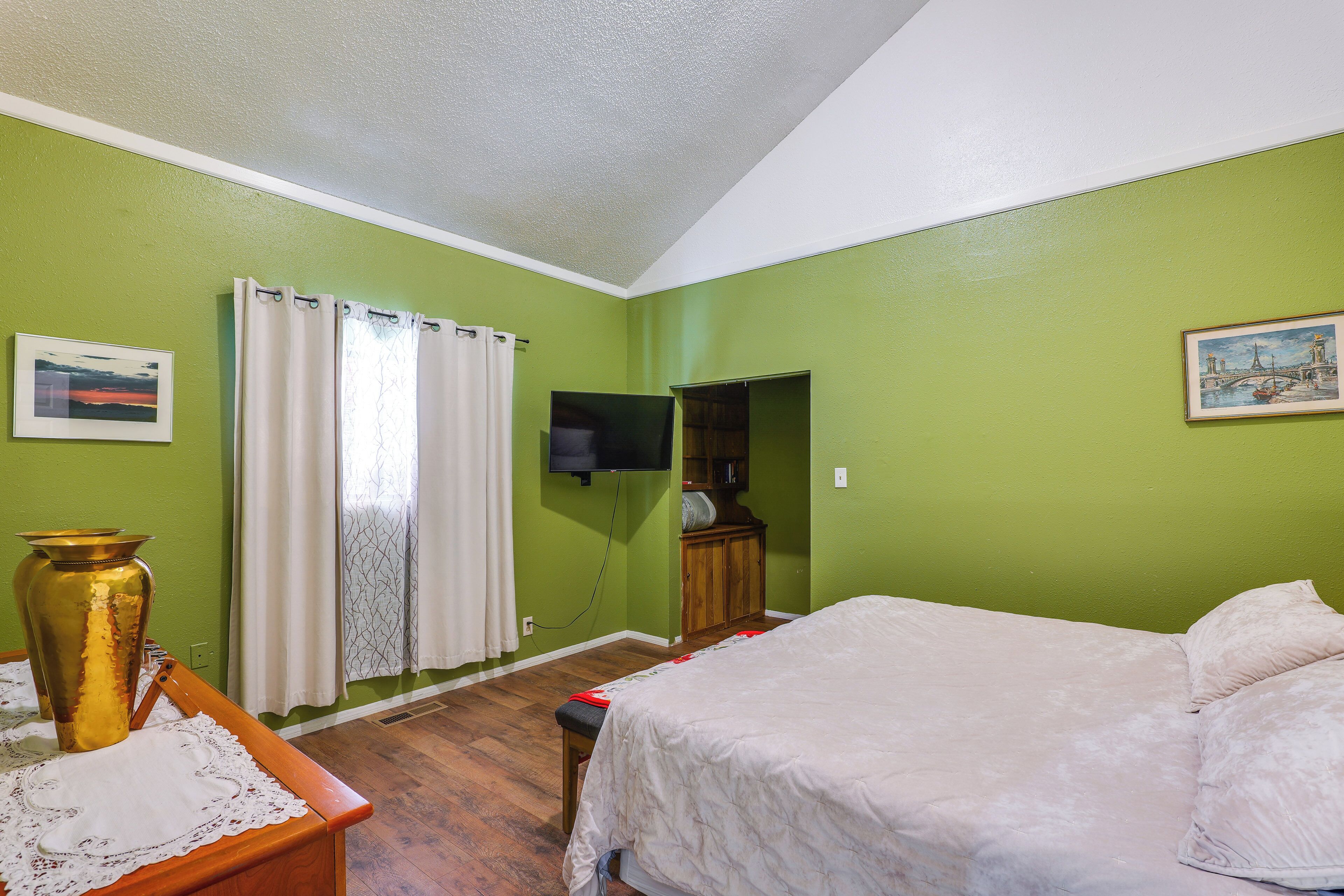A room with a bed, a television mounted on the wall, a window with curtains, and a framed picture on the wall.