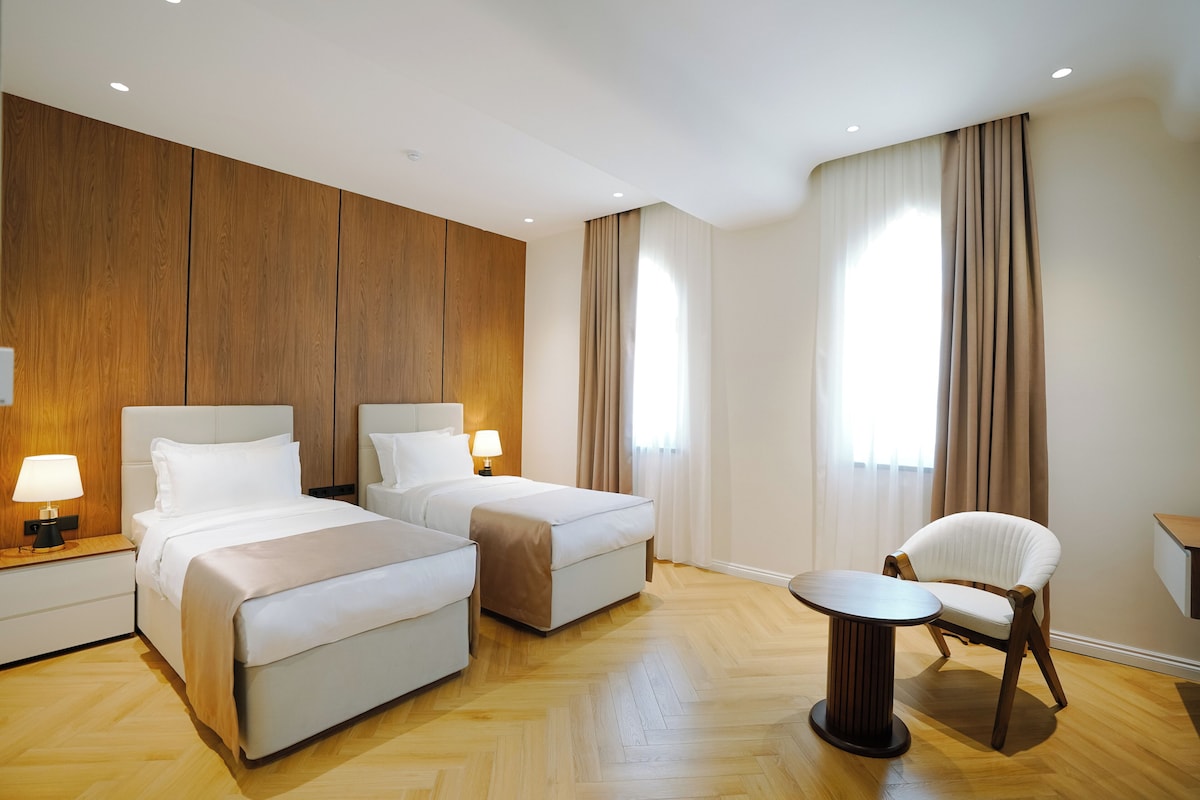 A hotel room with two beds, a chair, a small table, and a wooden accent wall.