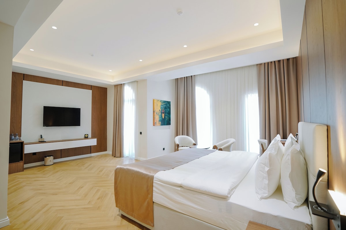 A modern hotel room with a large bed, a flat-screen TV mounted on the wall, a seating area with chairs, and a painting on the wall.