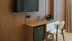 Habitación de hotel moderna con pared de paneles de madera, televisor de pantalla plana sobre un escritorio de madera, un minibar y una sola silla blanca.