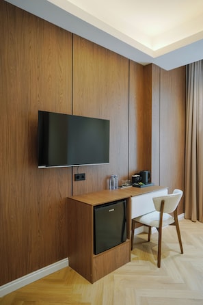 Habitación de hotel moderna con pared de paneles de madera, televisor de pantalla plana sobre un escritorio de madera, un minibar y una sola silla blanca.