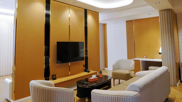 Una habitación de hotel moderna con televisión de pantalla plana montada en una pared de paneles de madera, un sofá, dos sillones, una mesita de centro y un escritorio con lámpara.