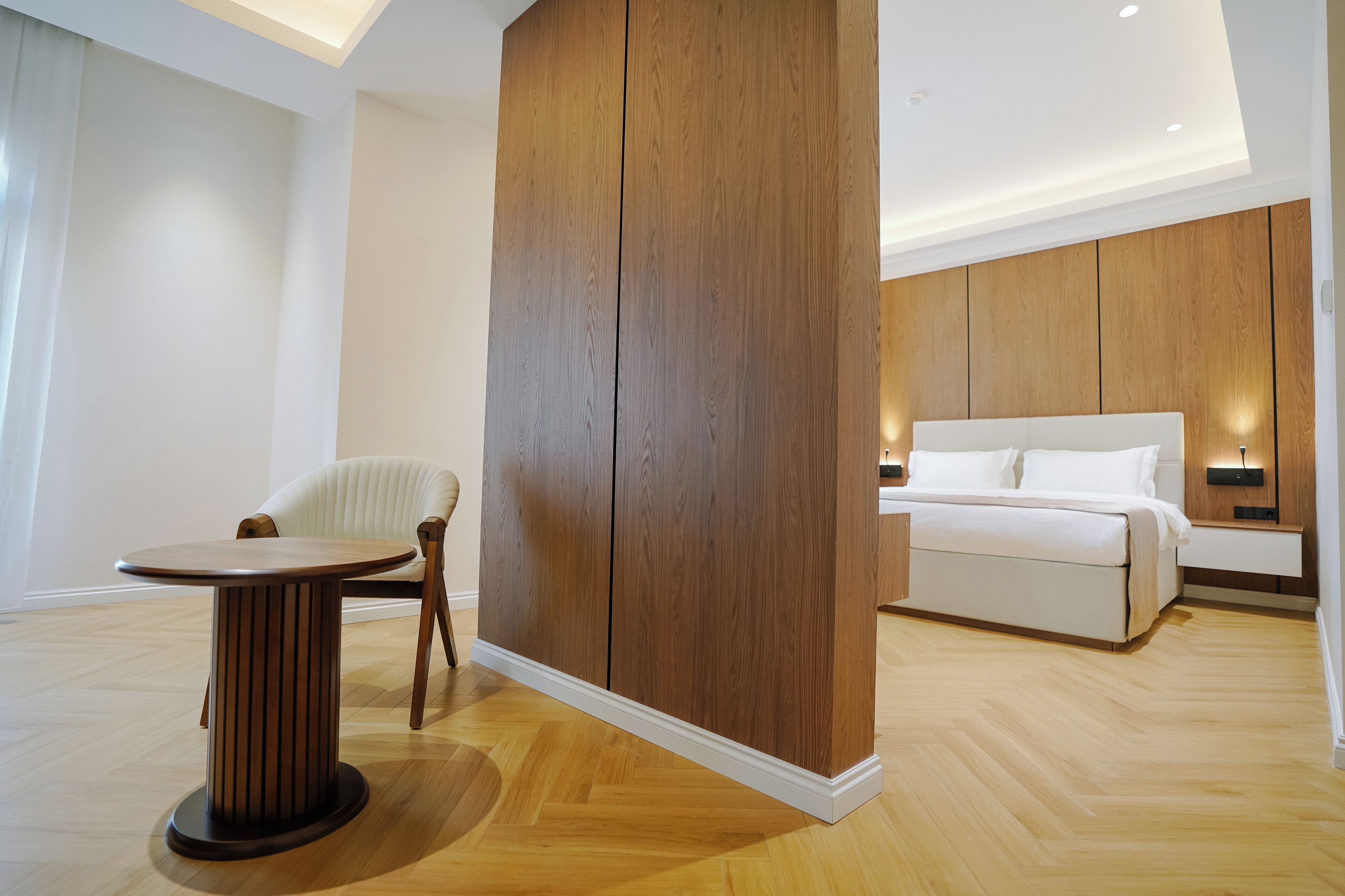 Ein modernes Hotelzimmer mit Holzboden, einem runden Tisch, einem weißen Bett und einer großen Wandpaneele aus Holz.