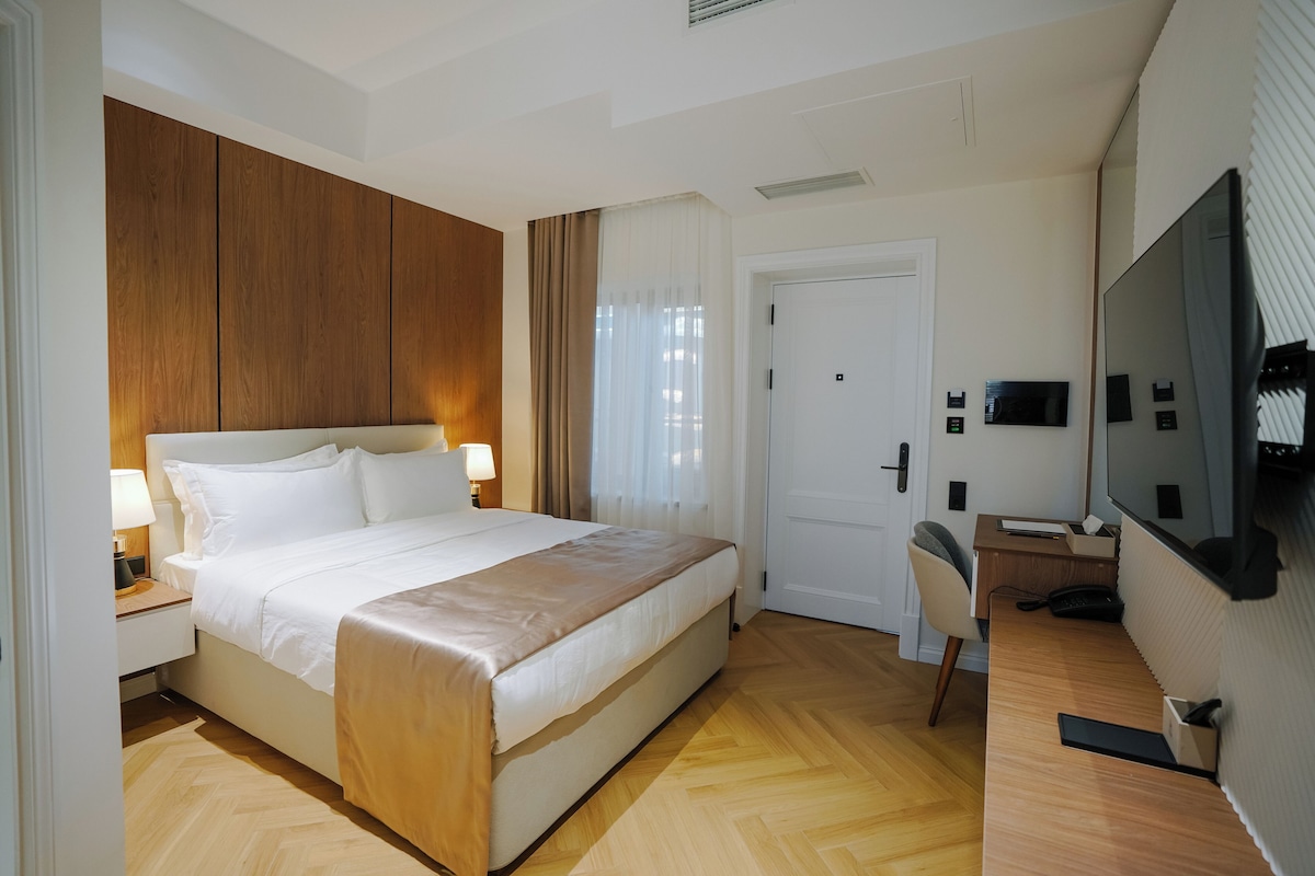 A hotel room with a bed, bedside lamps, a desk with a chair, a television, and a door.