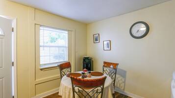 A dining area with a round table, two chairs, a clock on the wall, and a window with blinds.