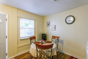 A dining area with a round table, two chairs, a clock on the wall, and a window with blinds.