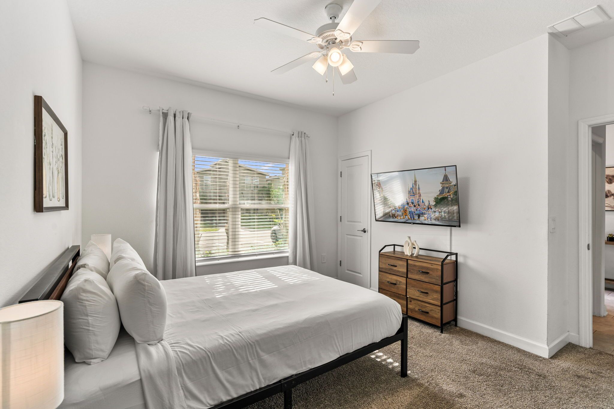 Une chambre à coucher avec un lit, une commode, un ventilateur de plafond, une fenêtre avec des rideaux et un téléviseur fixé au mur.