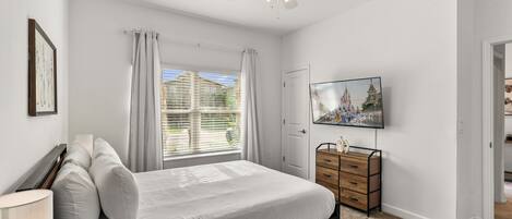 Une chambre à coucher avec un lit, une commode, un ventilateur de plafond, une fenêtre avec des rideaux et un téléviseur fixé au mur.