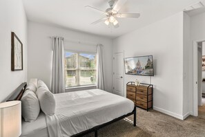 Ein Schlafzimmer mit einem Bett, einem Schminktisch, einem Deckenventilator, einem Fenster mit Vorhängen und einem Fernseher an der Wand.
