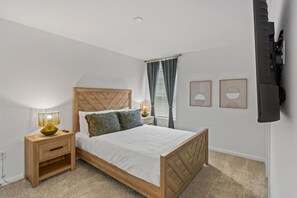 Ein Schlafzimmer mit einem hölzernen Bett, zwei Nachttischen, einer Lampe, einem Fenster mit Vorhängen und einem an der Wand befestigten Fernseher.