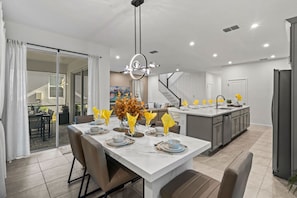 A modern kitchen with a central island, a dining area with a white table set for four, and a staircase leading to an upper level. - Brand New 6BR Villa Pool Spa PS5 Loft 2380 (Kissimmee)