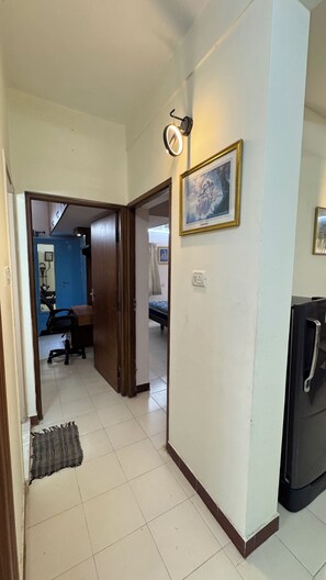 A hallway with a door leading to a room with a bed and a chair. There is a framed picture on the wall and a light fixture above the door. - Fully Furnished 2BHK in Anna Nagar Near Hospitals and IVF Centres (Chennai)