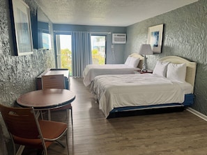 A hotel room with two beds, a table, a chair, and a window with curtains. - Ocean 12 Beach Hotel (Myrtle Beach)
