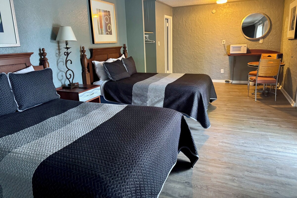 A hotel room with two beds, a desk with a chair, a lamp, and a framed picture on the wall.