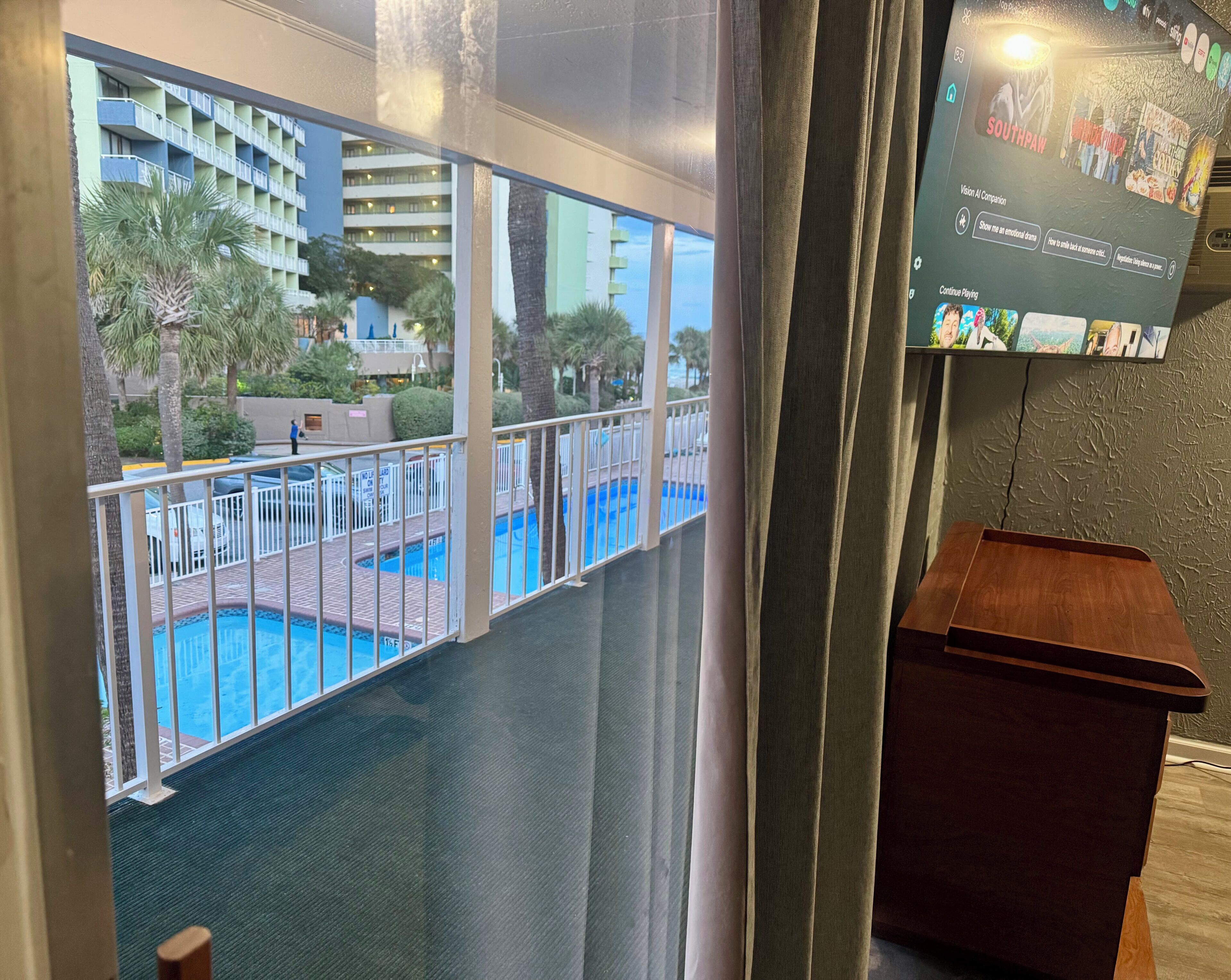 Uma varanda com piscina, vista de um edifício de hotel e uma televisão exibindo um programa.