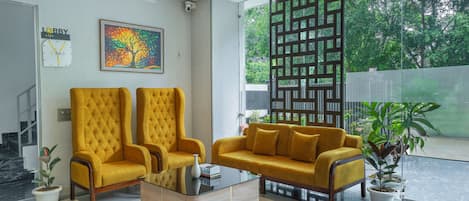 A modern lobby with two yellow armchairs, a coffee table, a painting, a clock, and a view of greenery outside.
