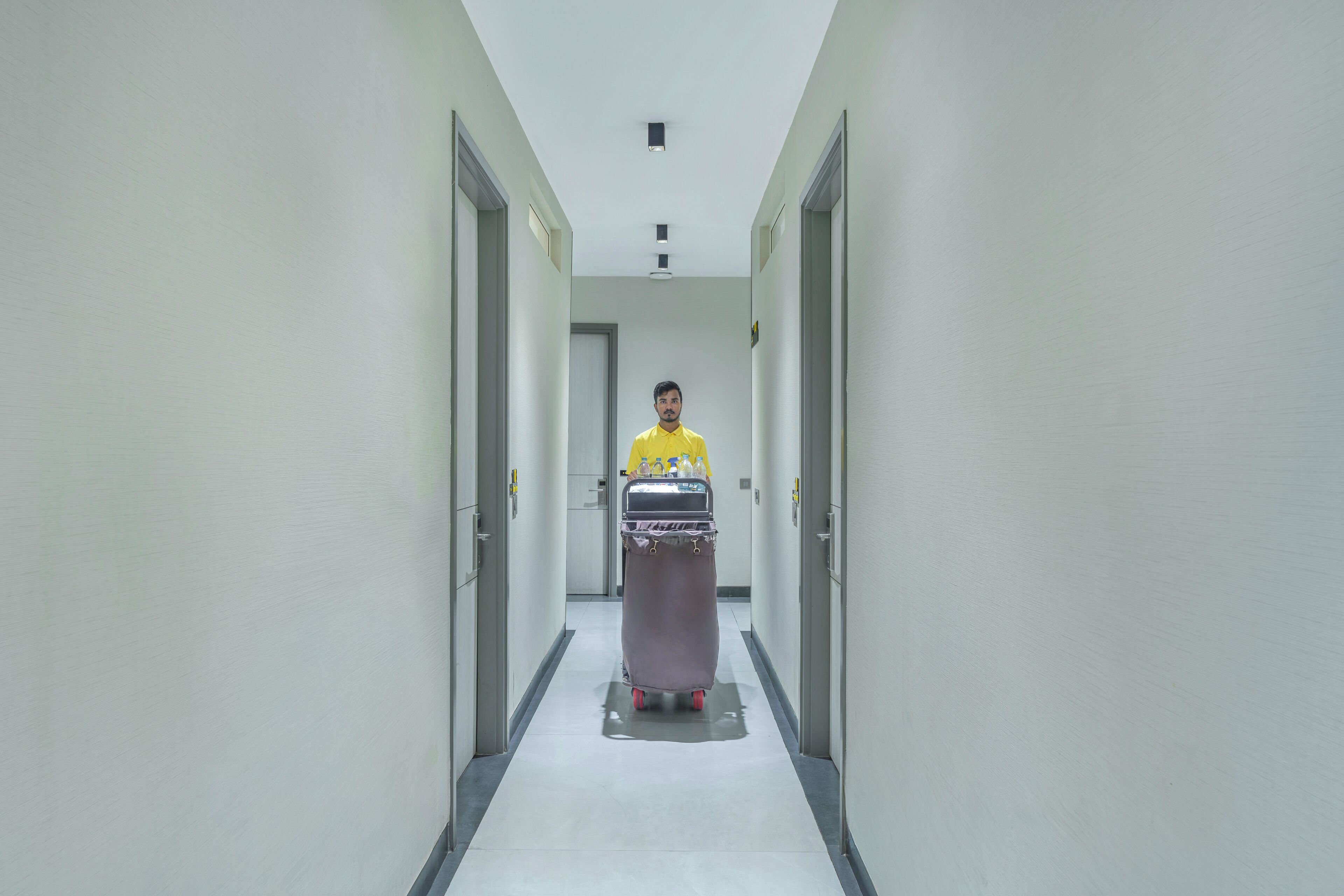 A person in a yellow shirt pushing a cart with water bottles down a hallway with doors on either side.