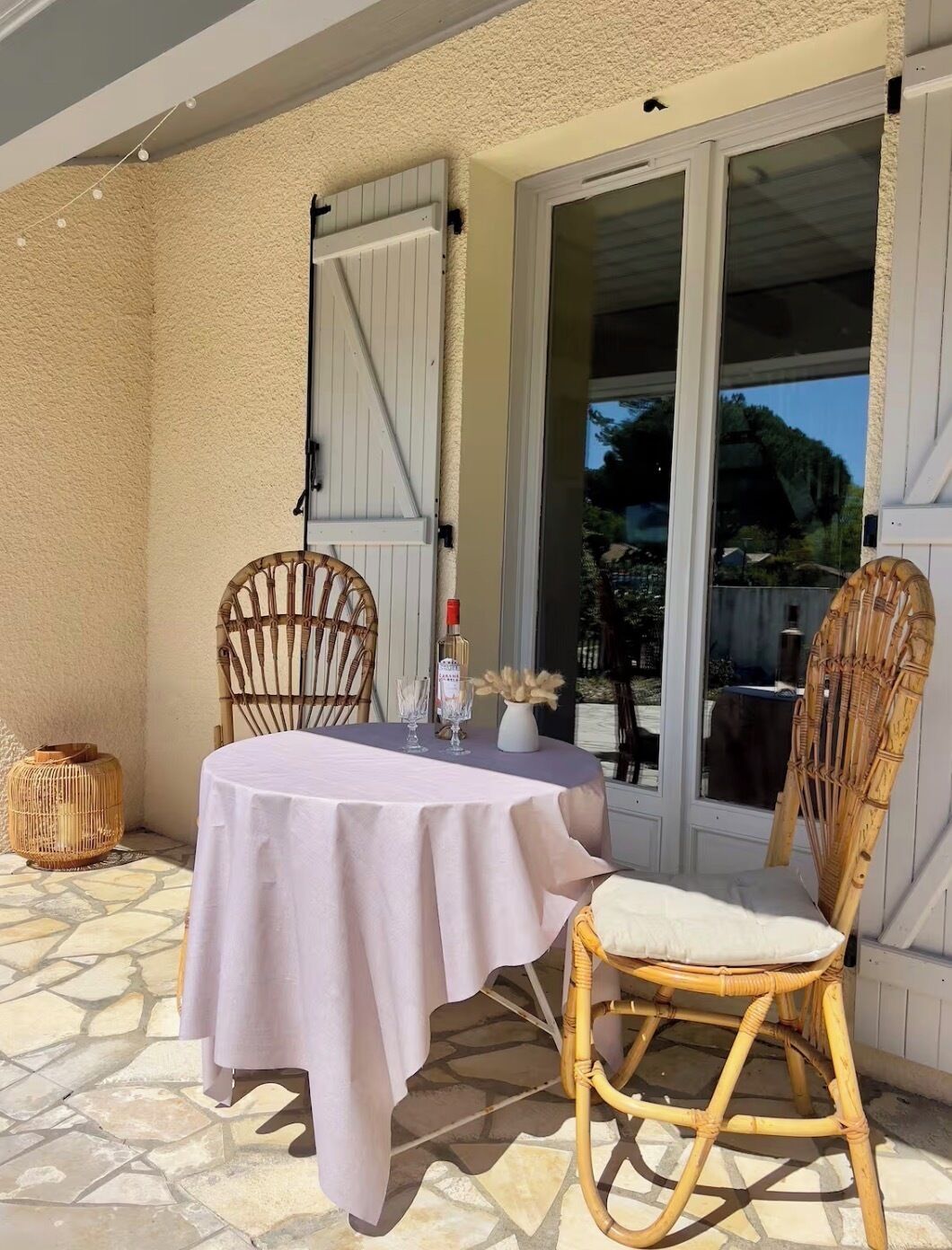 An outdoor patio with a round table, two wicker chairs, a vase with flowers, and a bottle.