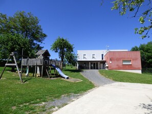 Un parque infantil con tobogán y estructuras para escalar, un edificio de fachada roja y un cielo despejado.