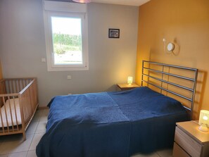 Un dormitorio con una cama, una cuna, una ventana y una mesita de noche con una lámpara.