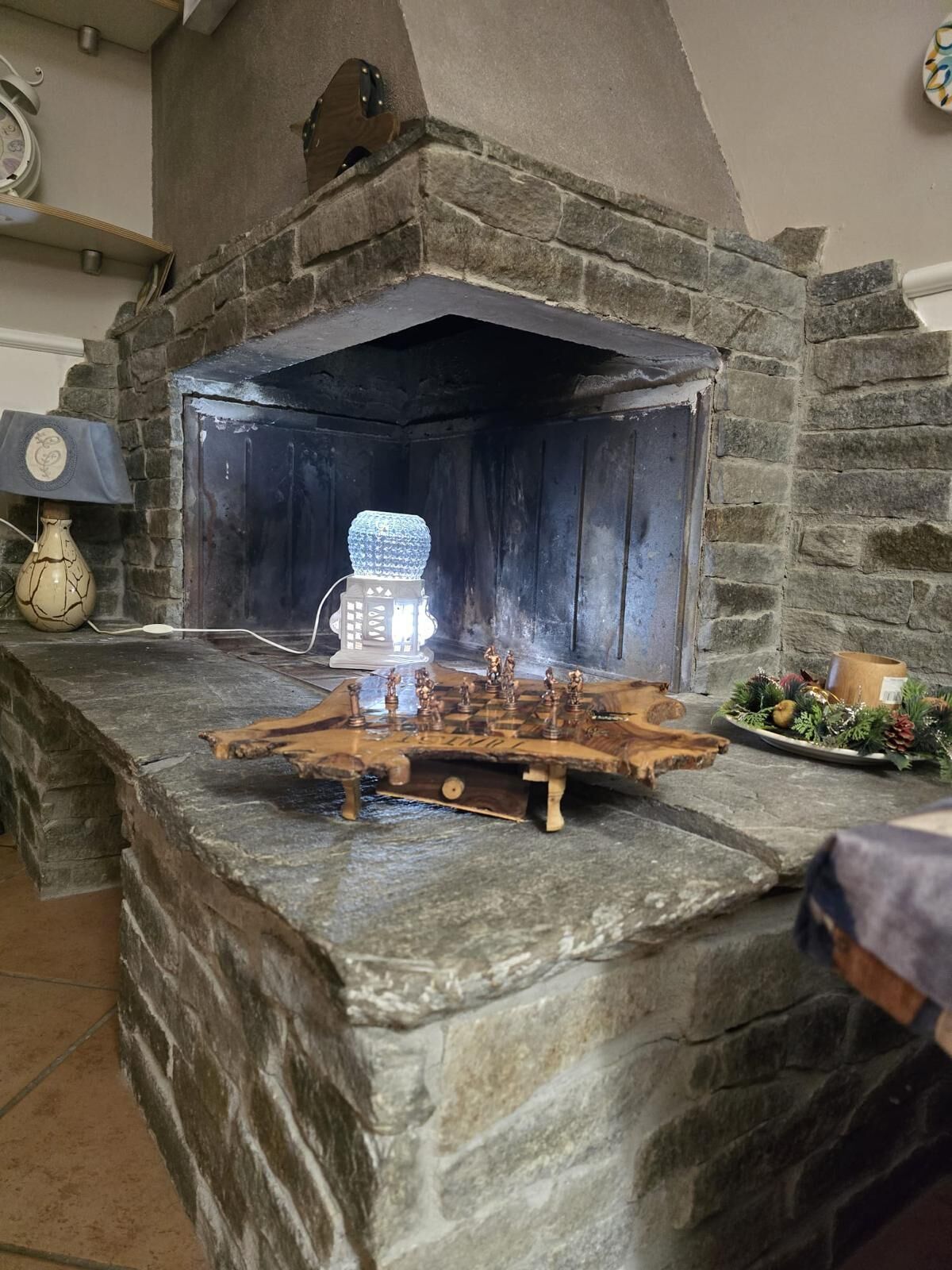 Une cheminée en pierre avec un poêle à bois intégré, un jeu d’échecs et une assiette décorative ornée de motifs végétaux.