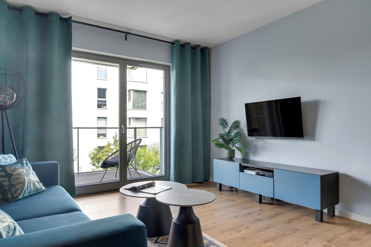 A modern living room with a sofa, a small round table, a TV mounted on the wall, and a balcony with a view of buildings.