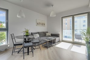 A modern living room with a sofa, a dining table with chairs, and a coffee table. There is a painting on the wall and a window with a view of buildings. - Nadmorze Seaside Apartment with Parking  (Gdańsk)