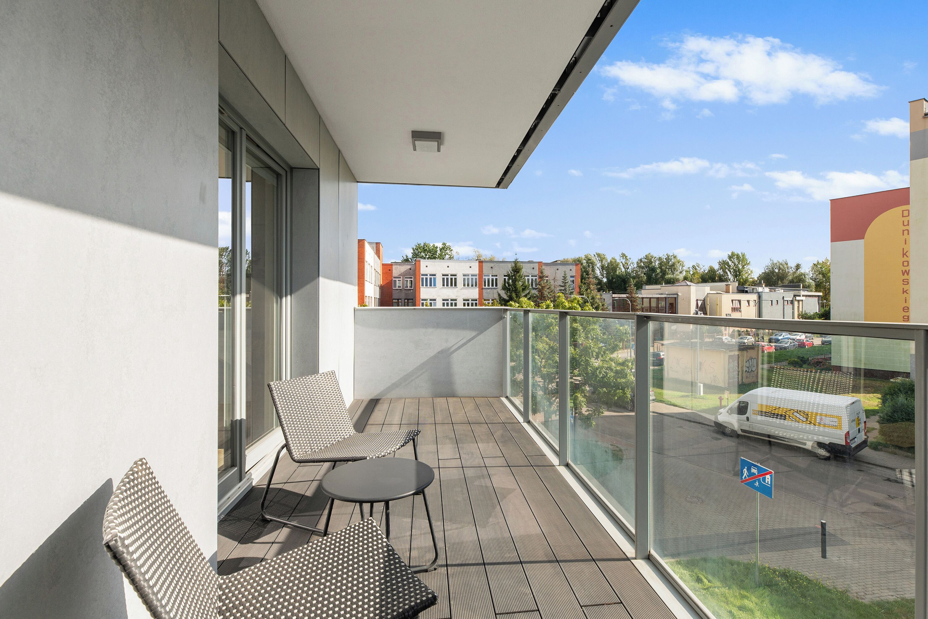 Un balcón con una silla, una mesa, barandilla de vidrio y vistas a edificios y una calle.