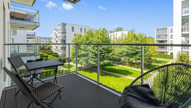 Varanda com guarda-corpo de vidro, mobiliário de vime e vista para edifícios vizinhos e vegetação.