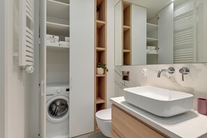 A modern bathroom with a built-in washing machine, a toilet, and a large mirror above a sink. - Nadmorze Seaside Apartment with Parking  (Gdańsk)