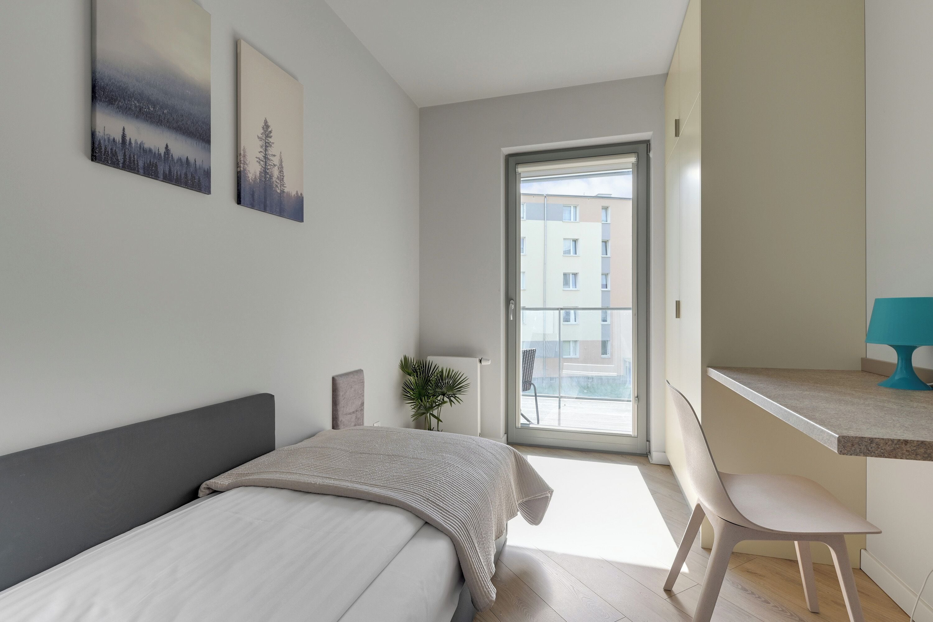 A modern bedroom with a bed, a desk, and a view of a building through a glass door.