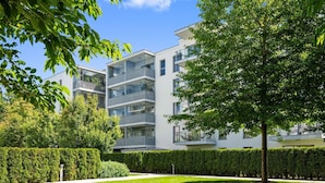 Edifício residencial moderno de vários andares com varandas, cercado por sebes bem cuidadas e um gramado.