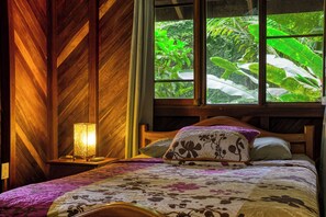 A bedroom with a wooden slat wall, a bed with floral bedding, a nightstand with a lamp, and a window with a view of greenery. - Wooden Cabin with Yoga deck over Creek, Walk to the Beach (Puntarenas Province)