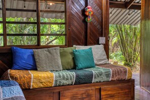 A wooden sofa with a quilted blanket and multiple cushions in a room with wooden walls and a window overlooking greenery.