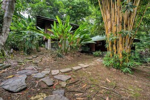 A garden with a stone pathway leading to a wooden structure surrounded by bamboo and tropical plants.