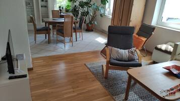Un salon comprenant un coin repas, un plancher en bois, une grande plante en pot, un meuble et un téléviseur.