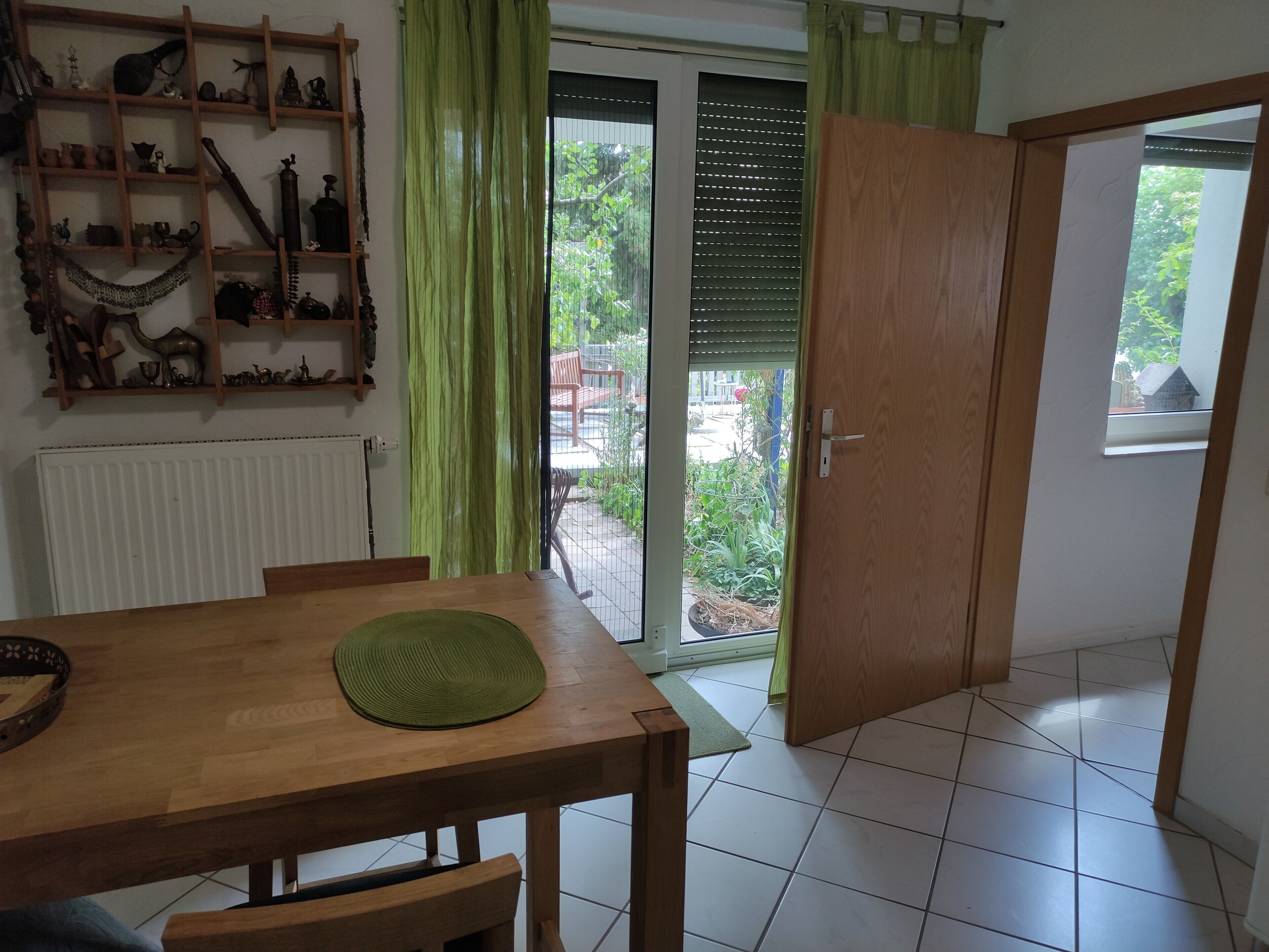A room with a wooden dining table, a green mat, a radiator, a sliding glass door to a garden, and a wooden door.
