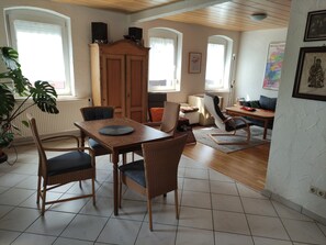 Uma área de refeições com mesa de madeira e cadeiras de vime, uma cozinha com armário e uma sala de estar com sofá e mesa de centro.