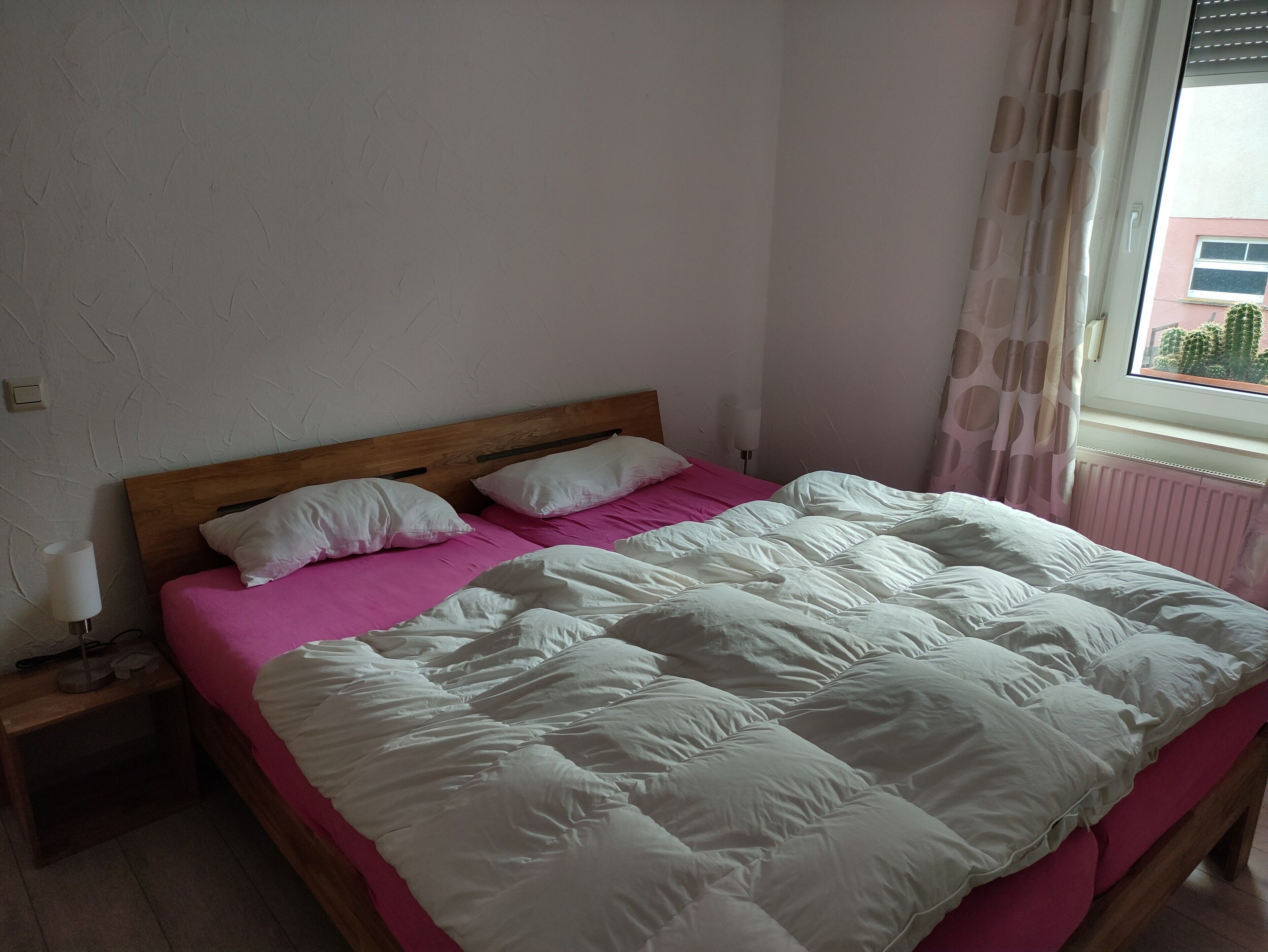 A bedroom with a wooden headboard, a bed with white and pink bedding, a bedside lamp, and a window with curtains.