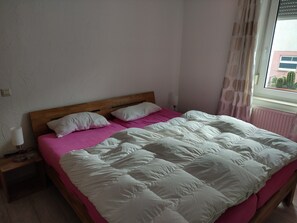 Quarto com cabeceira de madeira, cama com roupa de cama branca e rosa, abajur e janela com cortinas.
