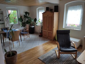 Ampla sala de estar com piso de madeira, mesa de jantar com cadeiras, uma poltrona preta estofada e vários vasos com plantas.