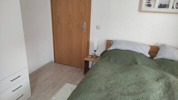 A bedroom with a wooden door, a bedside table with a lamp, a bed with green bedding, and a framed picture on the wall.