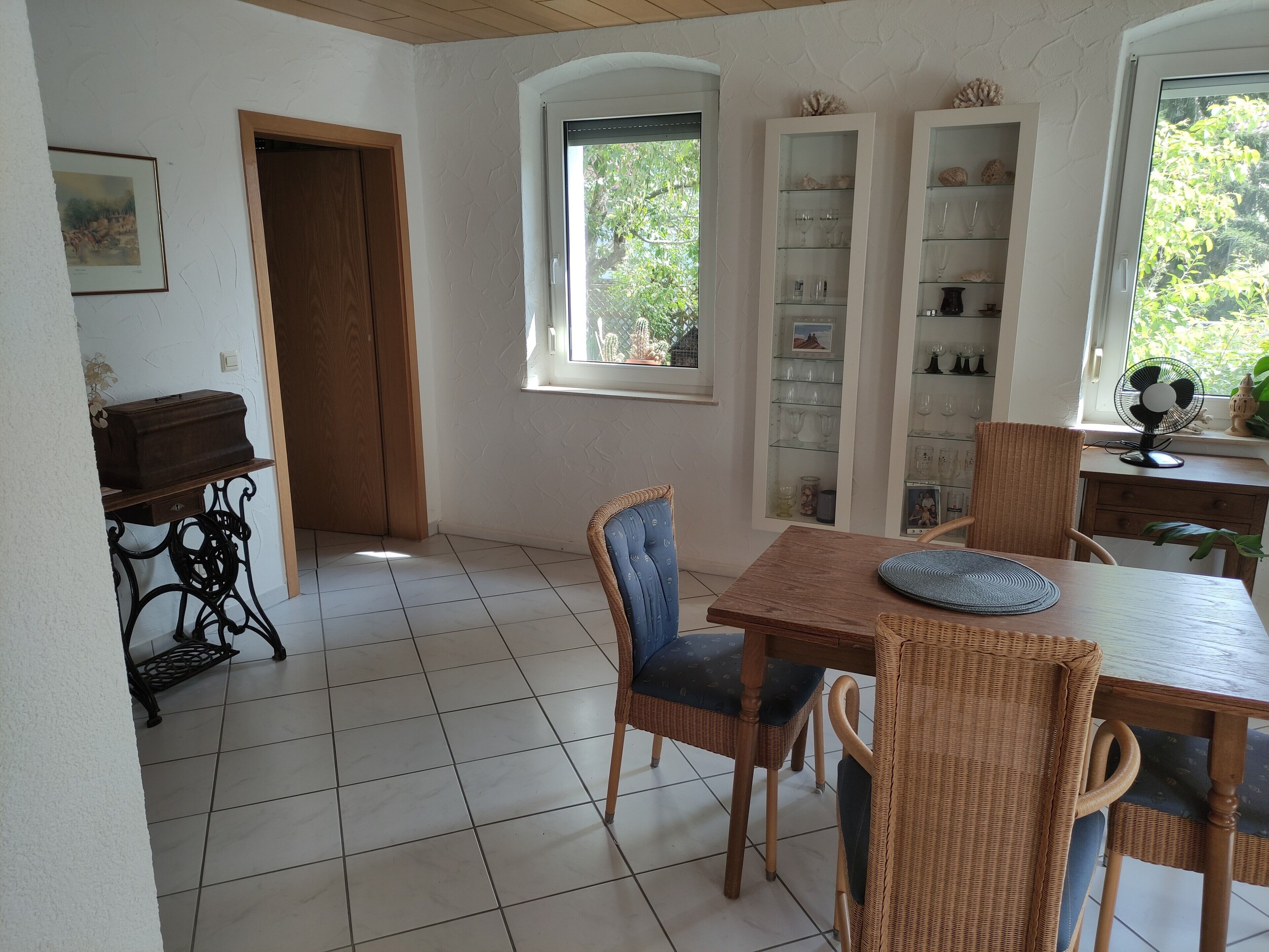 A dining area with a wooden table and wicker chairs, a white tiled floor, and a window with a view of greenery.
