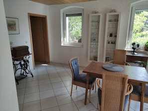 Un comedor con mesa de madera y sillas de mimbre, piso de baldosas blancas y ventana con vista a follaje.
