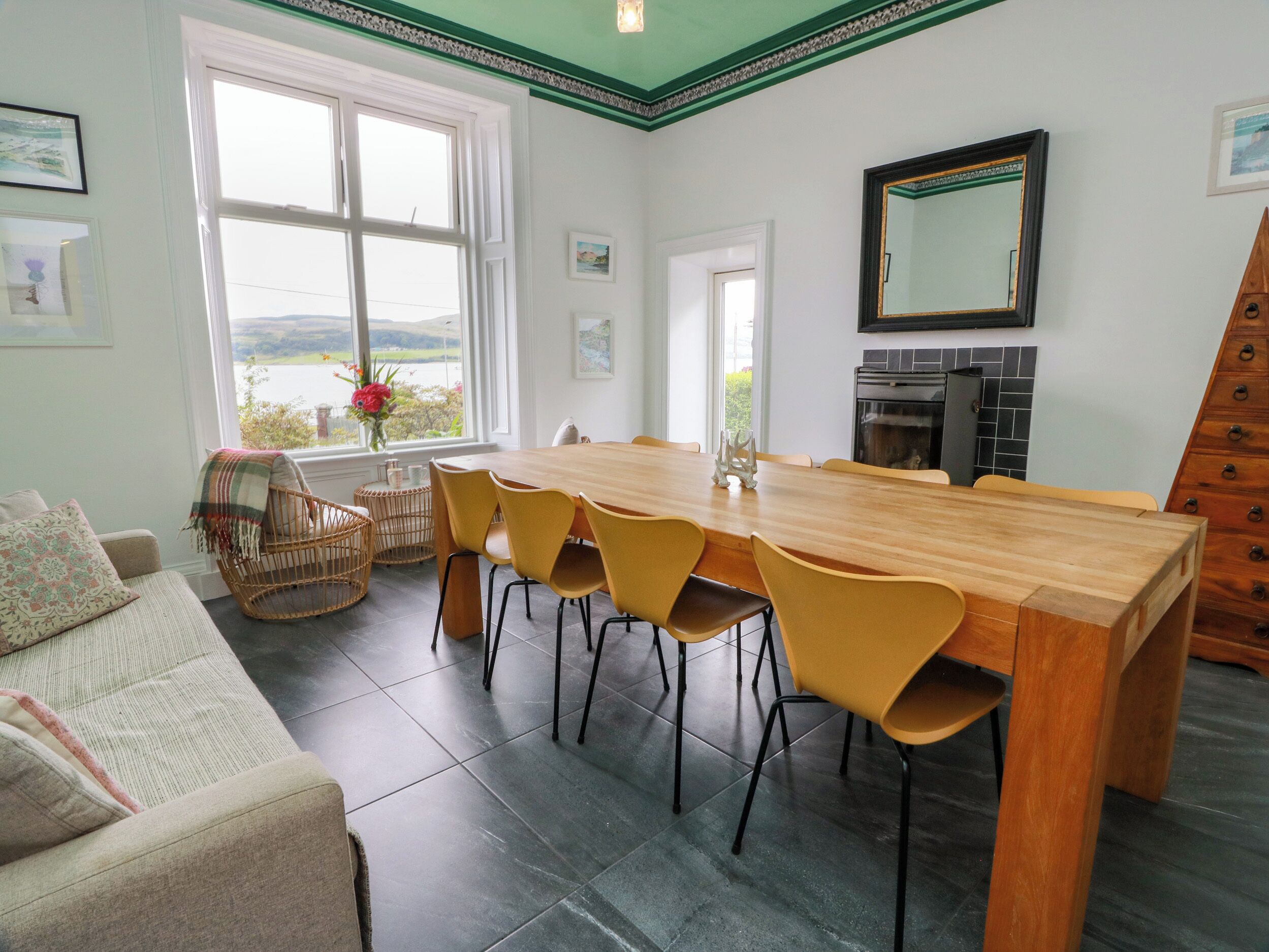 A dining area with a wooden table and yellow chairs, a wicker chair, a sofa with patterned cushions, and a view of the outdoors through a large window.
